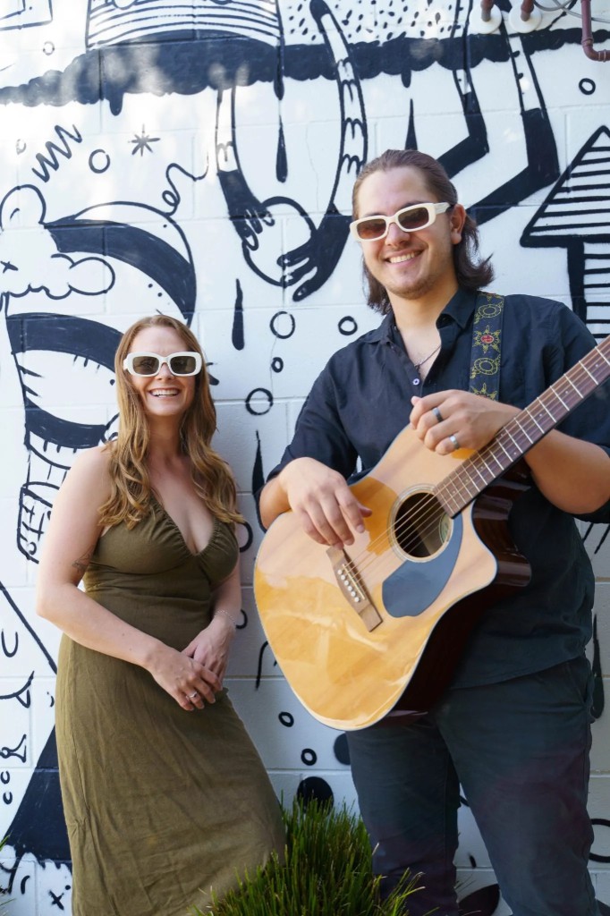 Sammie and Angelo with acoustic guitar in front of a mural